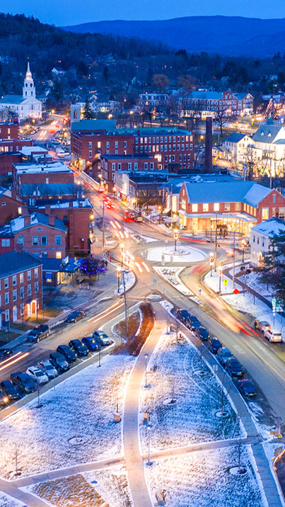 An aerial photo of downtown Middlbury, Vermont in the winter at night. Lights can be seen from cars and red brick buildings which line the street. In the foreground is a park with a light covering of snow.
