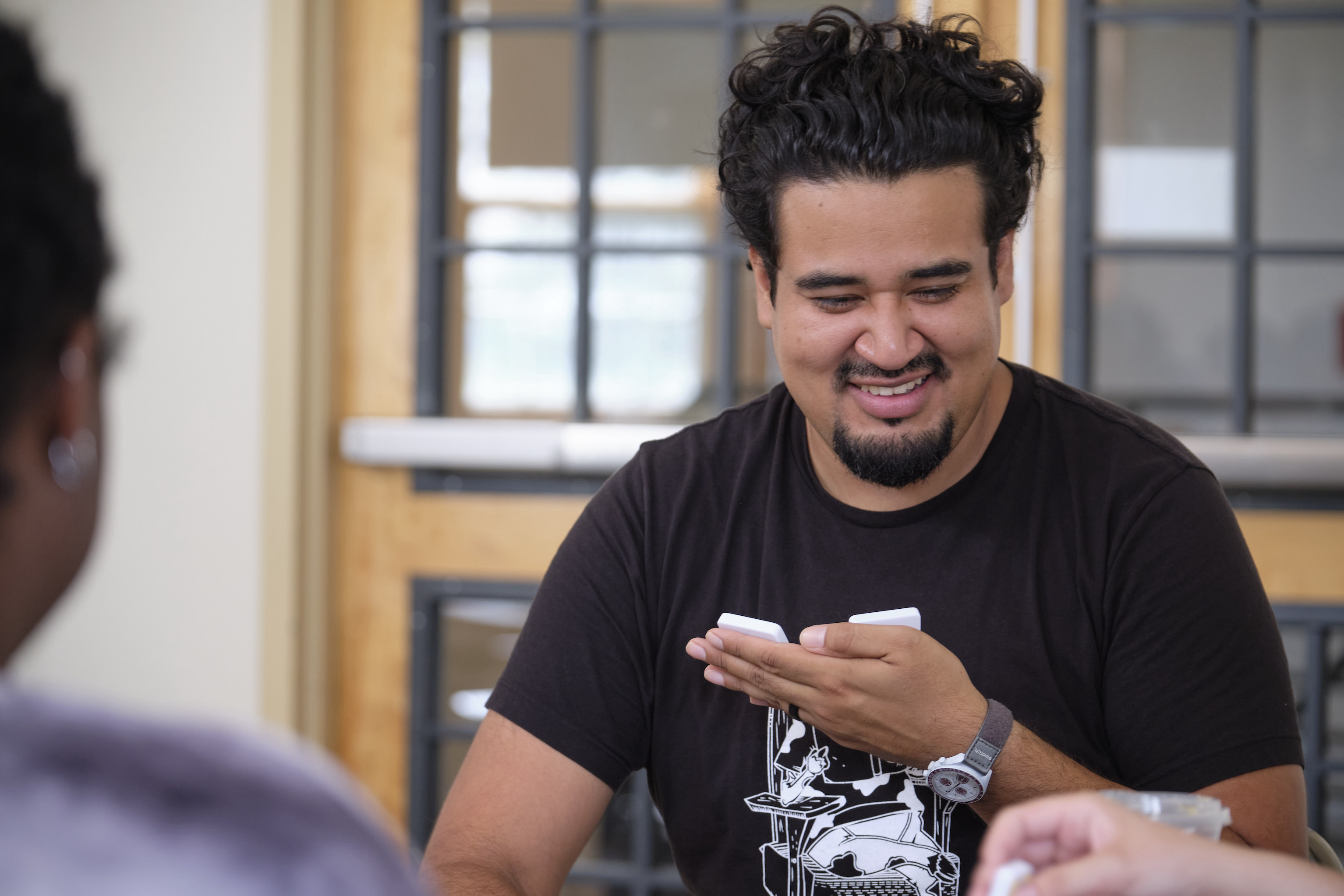 A young man smiles as he looks at the domino tiles in his hand.
