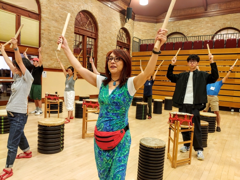 A group of people raise drum sticks into the air in unison, playing tall drums placed on the floor.