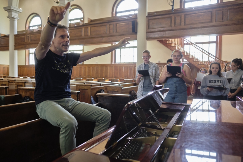 A man conducts a choir in a chapel. He sits behind a piano and gestures emphatically.