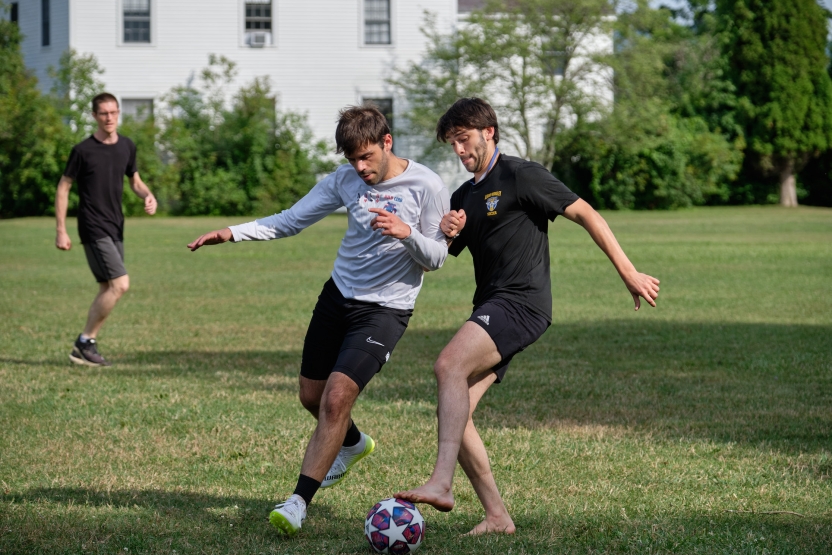 Two students play soccer outside in the grass.