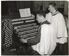 two men in church robes in front of an organ console, one seated at keyboard, the other turning pages of music