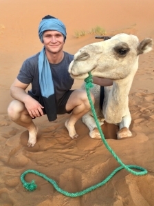 A student smiling in the sand next to a camel