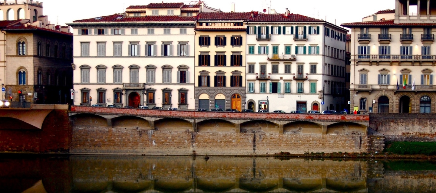 Old European-style apartment buildings line a street overlooking the Arno River in Florence.  The buildings' reflection are clearly shown in the river water.