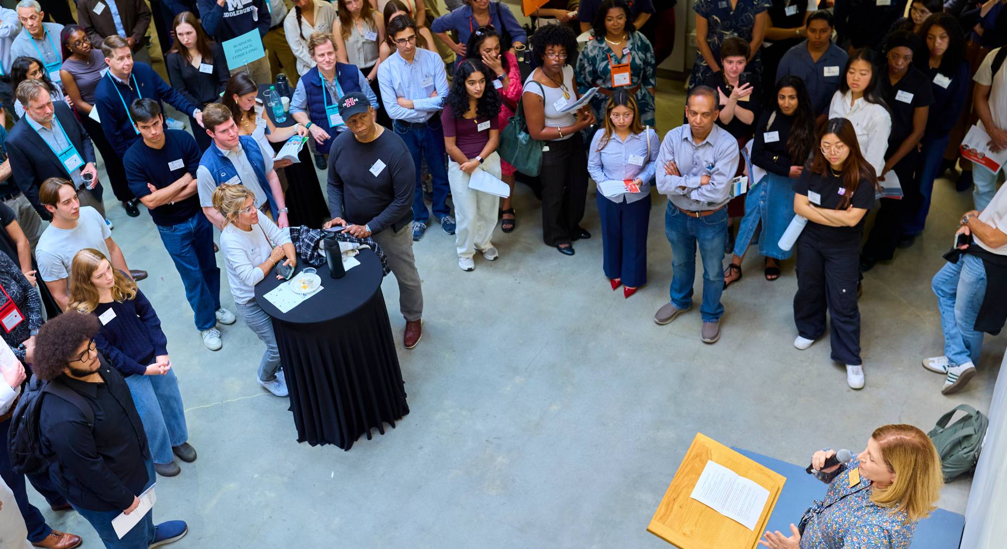 A photo of a large group of people standing around tables and listening to a speaker
