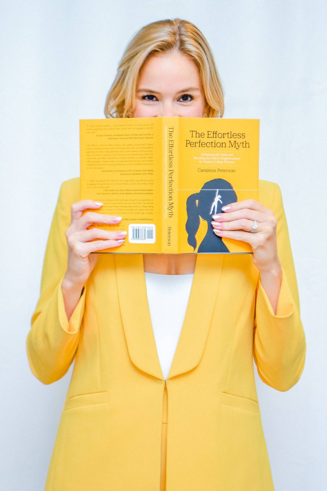 A woman with blond hair wearing a bright yellow suit and white jacket holds an open book in front of her face, the book jacket color matching the yellow of her suit. Her eyes appear to be smiling.
