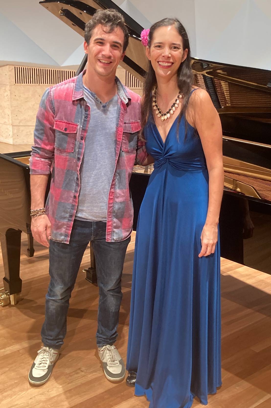 A white man with grey jeans, a grey henley shirt and a red and grey plaid button-down, stands in front of a grand piano beside a woman with long brown hair, a red flower in her hair, and a blue full-length satin gown. They are both smiling.