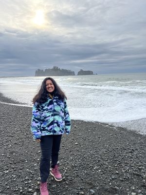Shivapriya Nair standing on a pebble beach wearing a jacket, pants and pink sneakers, smiling at the camera.