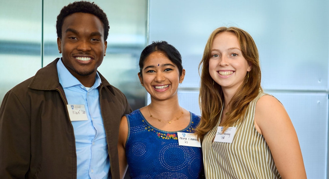 Photo of three students. From the left: a young man with short, black hair, a blue button-down shirt and a brown jacket, smiles into the camera and stands next to a young woman with a blue patterend tank top. She has dark brown hair, pulled into a pony tail. She is smiling into the camera. Next to her is a young woman is a green and white vertical striped tank top. She has long, redish-brown hair and she is smiling into the camera. They are all wearing name tags.