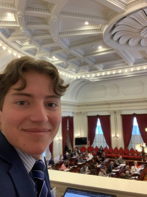 A young man faces the camera to take a selfie on the Vermont State House while the legislature is in session. He is wearing a blue suit and white and blue striped button-down shirt and blue tie.