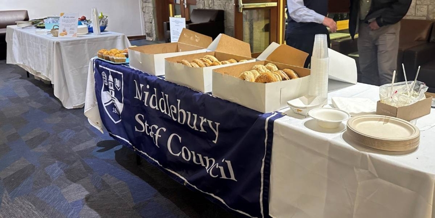 A photo of two catering tables with white tablecloths. The food served are bagels, muffins, doughnuts, fruit plate, and vegetable plate. There is a banner that reads, "Middlebury Staff Council"