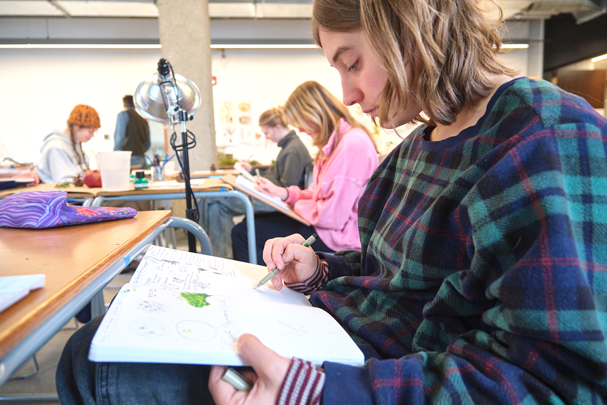 A student illustrates in a sketchbook at a classroom table.