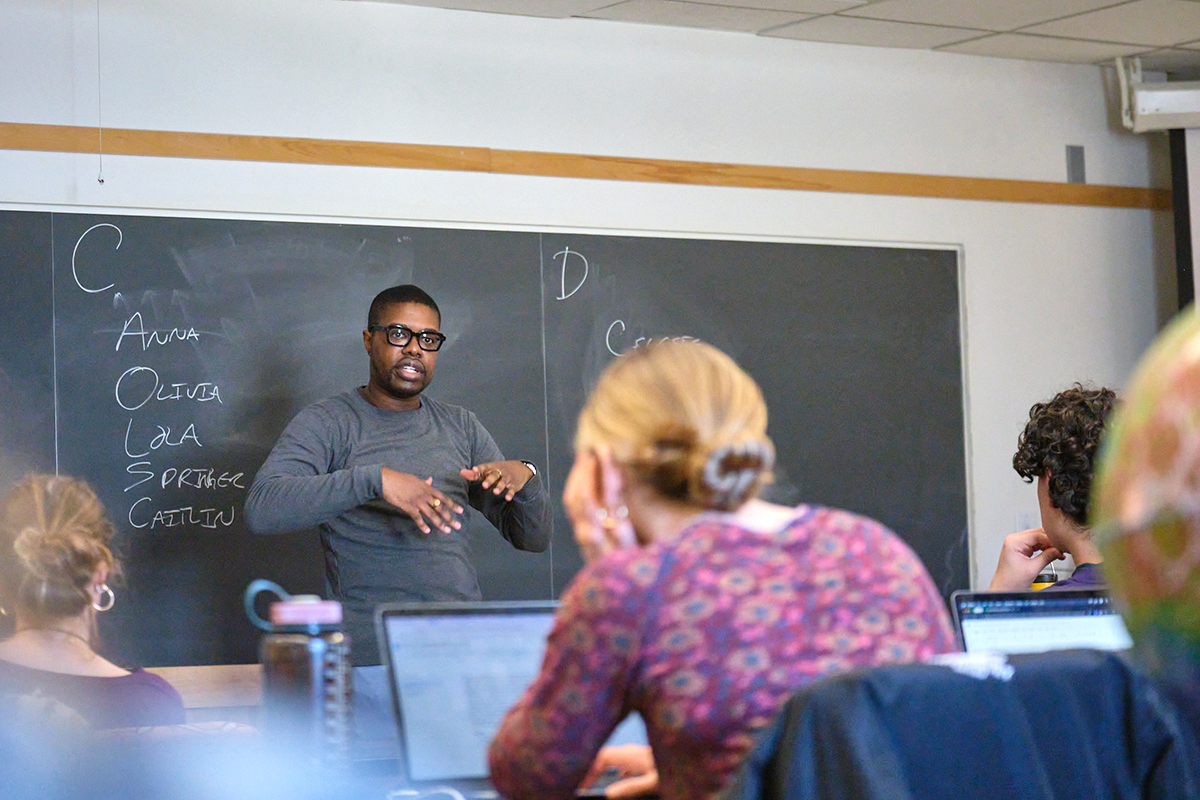 A professor lectures in front of a blackboard to a class of students.