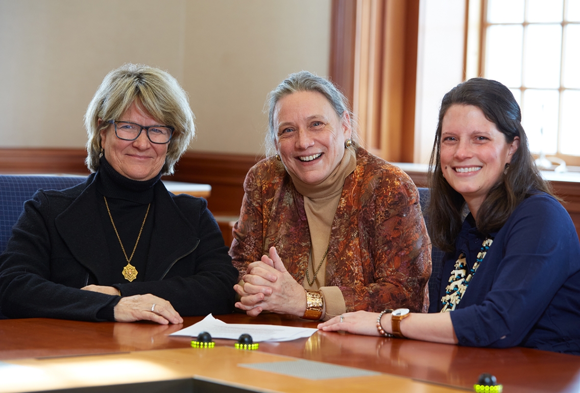 Photo shows three people sitting at a desk in a conference room.