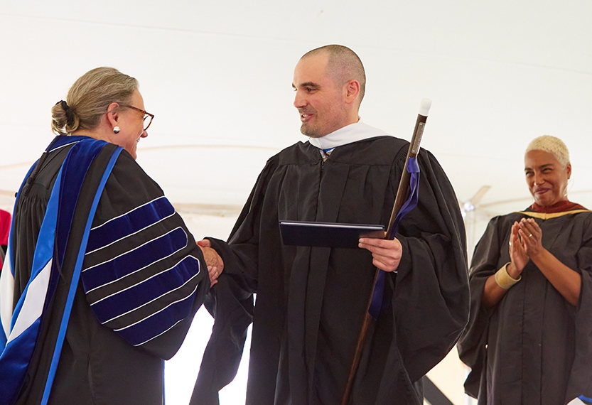Two people in academic regalia shake hands on a stage.