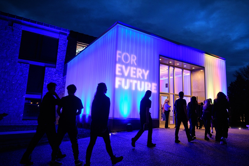 Students walk along a sidewalk in front of a lighted building at night.