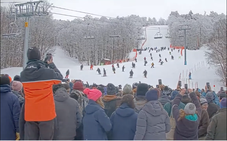 An audience watches graduates ski down the Middlebury College Snowbowl in academic regalia.
