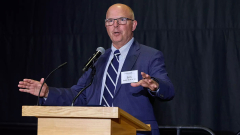 Man in dark blue suit and striped tie speaks at podium.