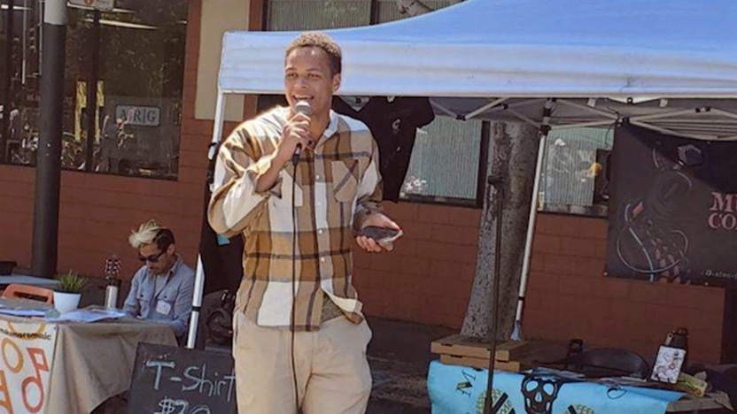 A college student stands on the sidewalk with a microphone in front of a tent.