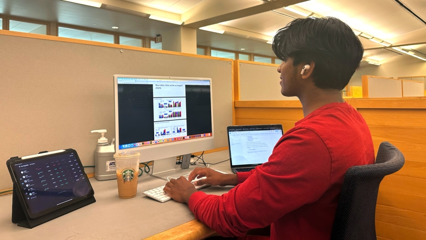 A student intern in a red shirt works at a computer station in an open office.