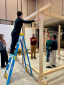 A student on a step ladder helps assemble a timber structure in a studio.