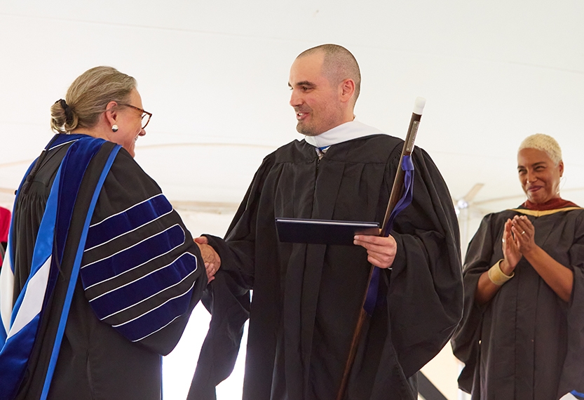 Two people in academic regalia shake hands on a stage.