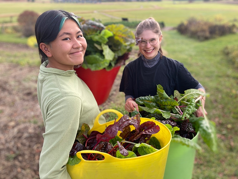 Two students stand in a garden with large buckets of produce.