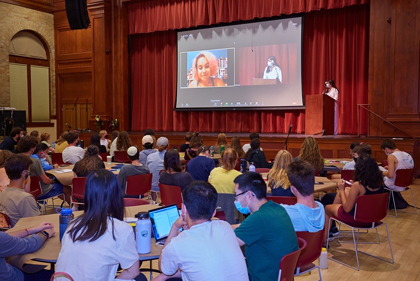 Students sitting at round tables in a large room watch a speaker on a projection screen.