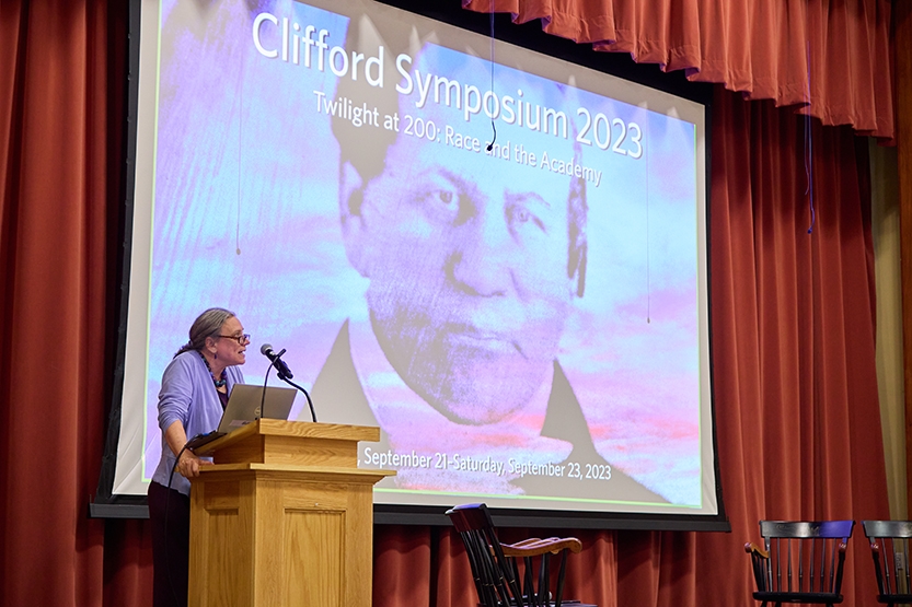 A speaker stands at a lectern onstage before a large projection screen.