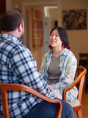 a student in a button-down over a Tshirt sits conversing with a professor wearing a black-and-white checked shirt, whose back faces the camera.