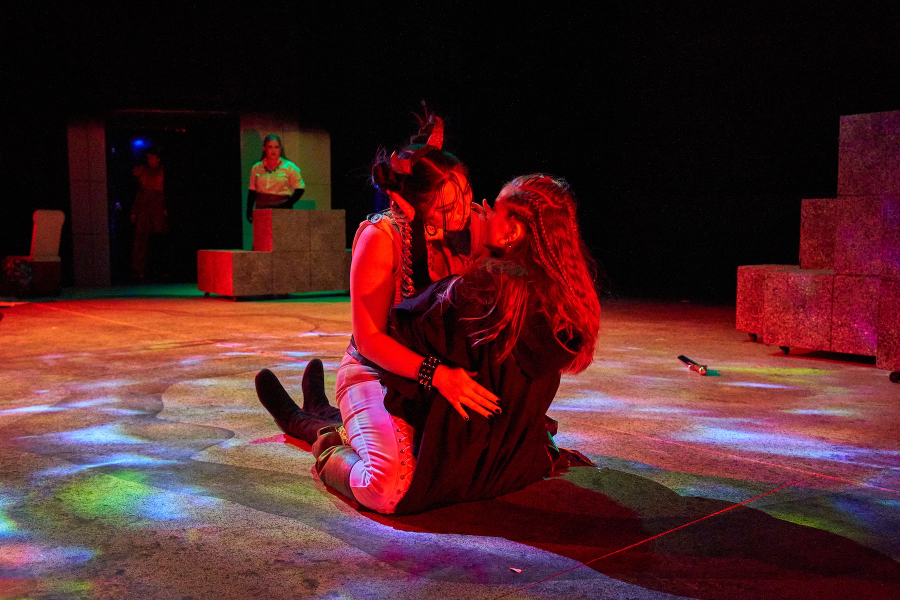 Two women kissing in red light as someone looks in the far background