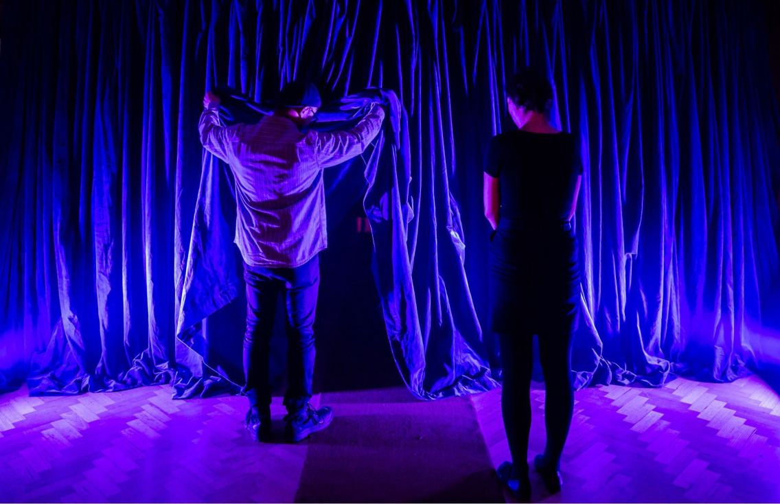 two people facing the curtain on a theatre stage. One is lifting the curtain up to see what is behind