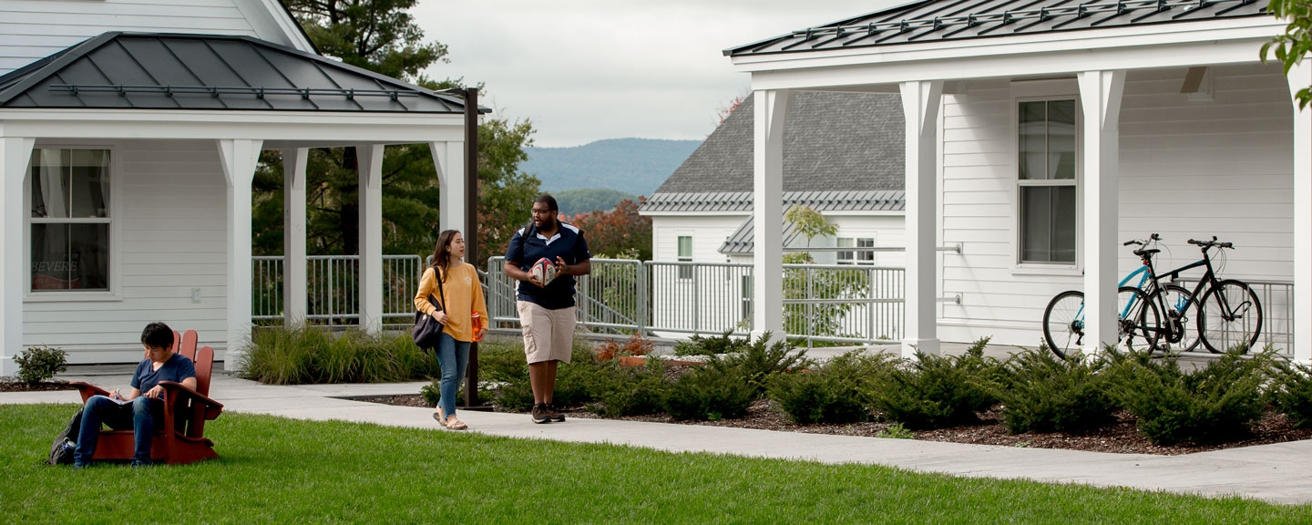 Two students walking in along a path in front of a dorm while another student reads in a chair on the lawn.