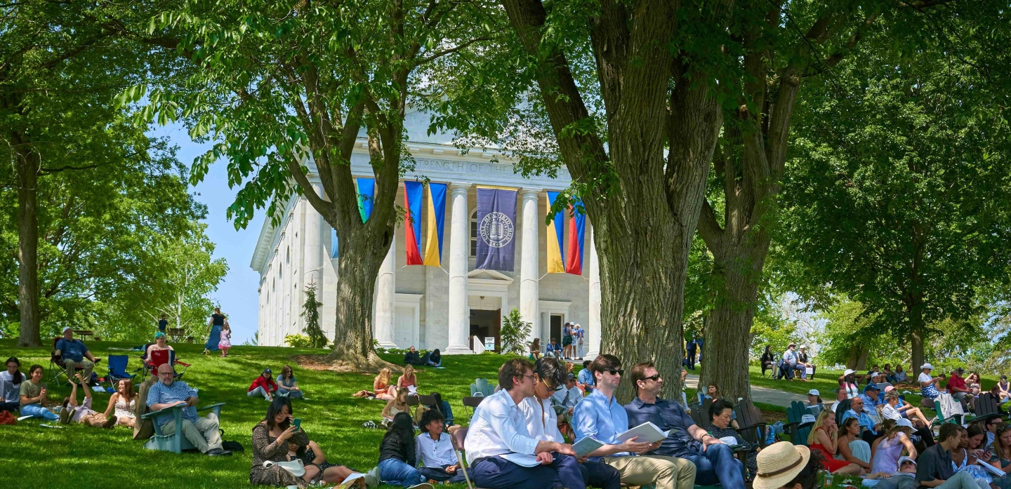 Middlebury Chapel on the hill with colorful commencement banners. Audience seated on the lawn beneath the trees.