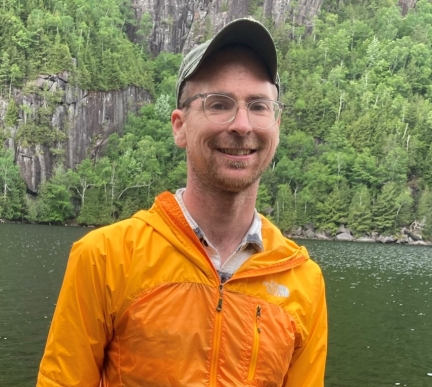 Man is bright orange jacket with ball cap and glasses standing by river