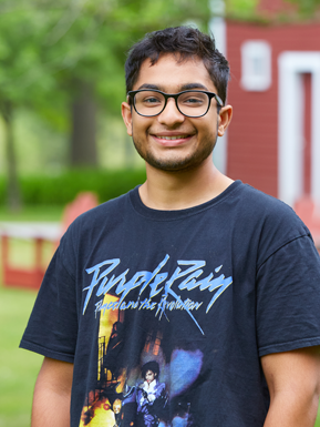 Photo of Vib Kumar wearing glasses and a Purple Rain t-shirt in front of a red building.