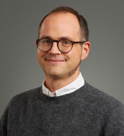 Man with short dark hair, dark rimmed glasses, grey sweater & whie collared shirt