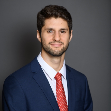 Photo of Germán Reyes. Person with dark hair and beard in a dark blue suit with red necktie on a grey background. 