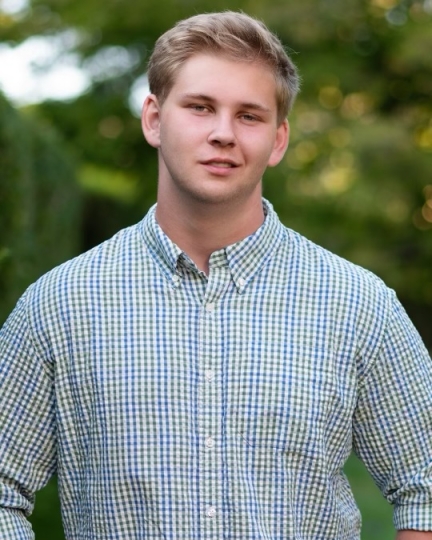 a photograph of Sam Spector, wearing a blue plaid button-down shirt and smiling at the camera