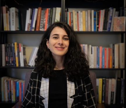 Fulya smiling at the camera with stacks of books in the background.
