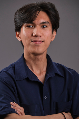 A photograph of Achilles Joshua, wearing a dark button-down shirt and smiling at the camera