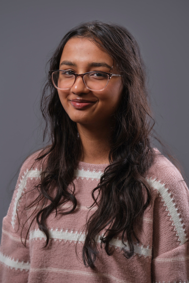 A photograph of Angabeen Mir, wearing a pink-and-white striped sweater and smiling at the camera