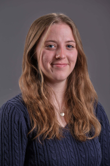 A photograph of Lucy Machon, wearing a blue sweater and smiling at the camera
