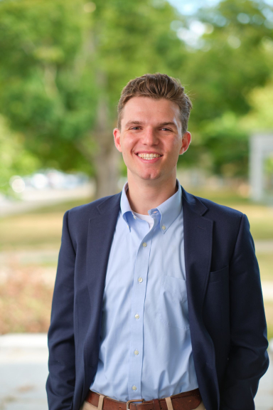 A photograph of Mack Briglin, wearing a blue button-down shirt and navy blazer, smiling at the camera