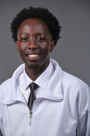 A photograph of Dan Tumwine, wearing a white quarter-zip over a white shirt and tie and smiling at the camera.