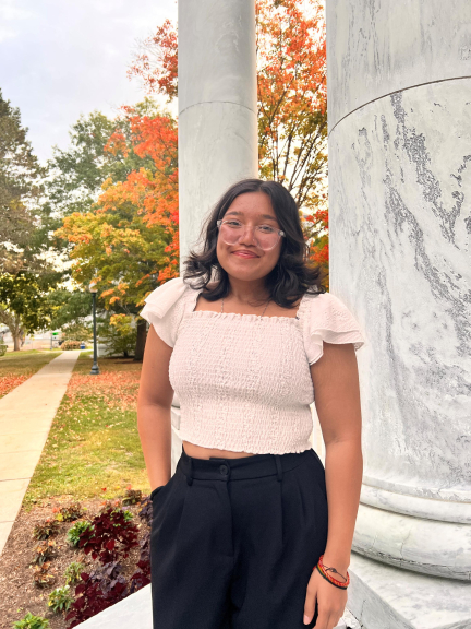 A photo of Munira Nurbhai, wearing a cream top and black pants, standing outside in the fall and smiling at the camera