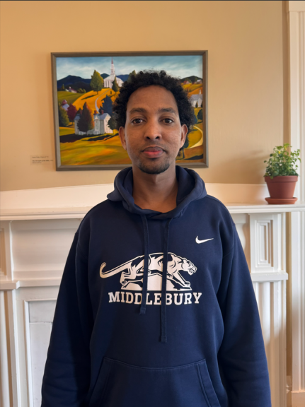 A photograph of Abdullahi Abdi standing in front of a fireplace with a painting of the Middlebury campus over the mantel, wearing a Middlebury sweatshirt