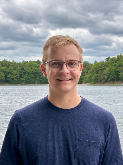 Headshot of Daniel Keliher who is smiling and wearing a navy blue shirt standing in front of a lake