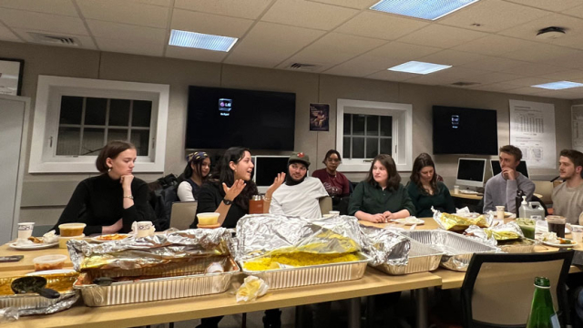 A student discusses her research while a large group is enjoying lunch at a table.