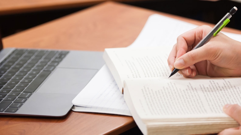 Close-up of a student writing in a book with a laptop.